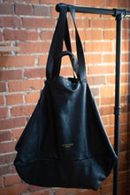 Load image into Gallery viewer, Black leather bag hanging on a stand against a brick wall.