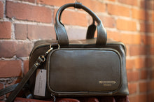 Load image into Gallery viewer, Brown leather handbag with a visible brand tag against a brick wall.