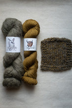 Load image into Gallery viewer, Two skeins of yarn in brown and gold with a knitted swatch on a light gray background.