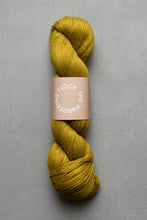 Load image into Gallery viewer, Mustard yellow skein of yarn with a label on a gray background