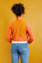 Load image into Gallery viewer, Person wearing a mustard yellow vest over an orange long-sleeve shirt with blue jeans on a yellow background