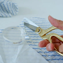 Load image into Gallery viewer, Gold-handled scissors held by a hand on a white and blue striped fabric background