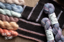 Load image into Gallery viewer, A styled flat-lay of yarn skeins and a knitted scarf on a textured beige fabric background. The yarn includes pastel blue, pink, peach, and lavender variegates, as well as a dark charcoal/black stripe pattern.