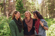 Three women standing together in a forest, smiling and wearing scarves.