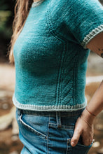 Load image into Gallery viewer, A close-up lifestyle shot of a person wearing a teal short-sleeve knit sweater with ribbed trim and blue jeans, standing outdoors with a blurred natural background.
