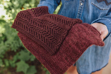 Load image into Gallery viewer, Person holding a folded maroon knitted garment with a blurred green background