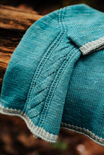 Load image into Gallery viewer, A close-up of a teal knitted textile, likely a hat or scarf, featuring cable knit and diagonal stitch patterns with a contrasting light gray ribbed trim, resting on a rustic wooden surface.