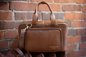 Brown leather bag with a smaller attached pouch against a brick wall.