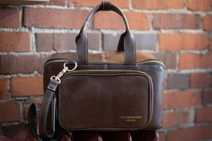 Brown leather handbag with a strap held against a brick wall.
