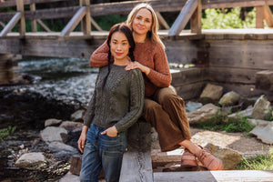 Two women sitting on a wooden bench by a stream with a wooden bridge in the background.