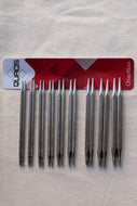 Ten metal knitting needles arranged in two rows of five on a light fabric background, with a red and white ChiaoGoo QUADS card placed behind them.