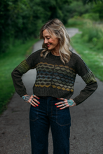 Load image into Gallery viewer, A woman stands on a paved path outdoors wearing a dark knit sweater with horizontal wavy stripes in olive green, gray, and mustard yellow across the chest and sleeves. She is also wearing dark blue jeans, with her hands on her hips.