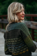 Load image into Gallery viewer, A woman with blonde hair is shown from the back wearing a dark knit sweater with green, blue, and yellow striped and patterned details, standing outdoors near a wooden railing.