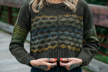 Load image into Gallery viewer, A person wearing a dark olive knit sweater with a colorful geometric pattern across the chest and green striped sleeves, standing outdoors on a wooden bridge.
