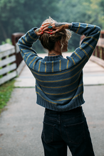 Load image into Gallery viewer, A person viewed from behind wearing a blue-gray ribbed knit sweater with thin mustard yellow stripes, standing on an outdoor path near a bridge.