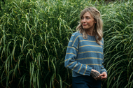 A woman with blonde hair stands outdoors in front of tall green grass, wearing a blue knit sweater with thin yellow stripes and dark pants.