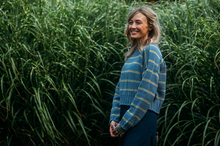 Load image into Gallery viewer, A woman with blonde hair stands in a field of tall green grass, wearing a blue knit sweater with thin yellow stripes and dark blue pants.