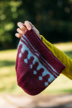Load image into Gallery viewer, A hand holding a maroon and light blue knit beanie with a Fair Isle pattern, set against a blurred green outdoor background.