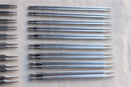 A set of metal knitting needles laid out on a light fabric background, with some needles grouped on the left side. The needles are silver-toned with printed size and brand markings.