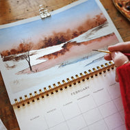 Calendar with a winter landscape illustration on a wooden surface, with a hand holding a pen.