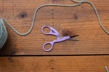 Load image into Gallery viewer, Lavender purple embroidery scissors with silver metal blades, resting on a rustic wooden surface with a ball of gray thread partially visible on the left.