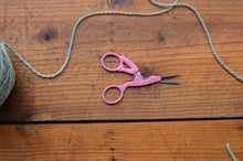 Load image into Gallery viewer, Pink-handled scissors with silver metal blades resting on a rustic wooden surface, with gray yarn draped across the scene and part of a ball of yarn visible at the left edge.