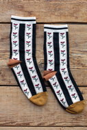 A pair of crew socks with black and white vertical stripes, decorated with small red rose motifs, brown heels, and mustard yellow toes, displayed flat on a rustic wooden background.