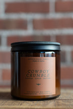 Load image into Gallery viewer, A soy wax candle in an amber glass jar with a black metal lid and a tan label reading 'COWBOY CRUMBLE' by Seventh House Candle, set against a blurred brick wall background.