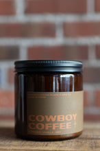 Load image into Gallery viewer, An amber glass jar candle with a black metal lid and a tan paper label reading 'COWBOY COFFEE' in orange, set on a wooden surface with a blurred brick wall background.
