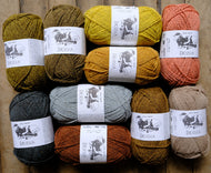 Ten skeins of Brusca yarn in assorted earthy colors, arranged in a 3x3 grid on a rustic wooden background. The colors include olive green, mustard yellow, terracotta orange, charcoal gray, light blue-gray, rust brown, ochre brown, and sandy beige.
