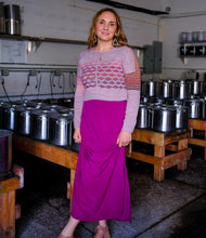 Load image into Gallery viewer, A woman stands in a kitchen or brewery setting with large stainless steel pots on wooden tables, wearing a lavender and pink patterned knit sweater with striped sleeves and a bright magenta maxi skirt.