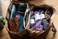 Brown leather bag filled with yarn, knitting supplies, and books on a wooden surface