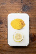 Load image into Gallery viewer, Two lemons, one whole and one sliced, on a white rectangular plate against a wooden background.