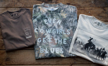 Load image into Gallery viewer, Three folded graphic t-shirts arranged on a rustic wooden surface. The left shirt is solid dark brown with a small white tag.