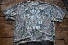 Load image into Gallery viewer, A short-sleeve crewneck t-shirt with a woodland camouflage pattern, featuring the text 'EVEN COWGIRLS GET THE BLUES' printed in white across the chest, laid flat on a rustic wooden plank surface.