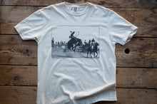 Load image into Gallery viewer, A white short-sleeve crewneck t-shirt laid flat on a rustic wooden plank background. The shirt features a black-and-white graphic print in the center depicting a cowboy riding a bucking bronco during a rodeo, with 'THE COWBOY CLUB' printed below the graphic.