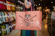 A person in a yarn shop holds up a peach-pink reusable tote bag with black text reading '1952 Farmer Daughter Fibers' and a ram skull graphic. The background shows shelves filled with colorful yarn skeins.