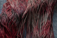 Load image into Gallery viewer, Close-up of variegated yarn strands in shades of deep red, burgundy, and gray, laid out on a textured gray fabric background.