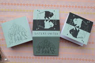Three square art cards or prints with black-and-white illustrated figures and the text 'SISTERS UNITED' are arranged on a pink patterned fabric background.