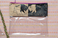 A clear zippered pouch with a fabric header that reads 'SISTERS UNITED' and shows two bison silhouettes, one on beige and one on black, laid flat on a patterned pink and orange surface.