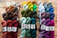 Three vertical groups of small yarn skeins in many different colors arranged on a rustic wooden surface. The left group includes warm neutrals, browns, grays, reds, and rust tones; the center group includes pale sage, olive, mustard, yellow, green, and blue-green; the right group includes light blue, gray, mauve, lavender, and bright purple.