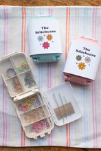 Load image into Gallery viewer, Three small storage cases labeled 'The Stitchcase' arranged on a striped fabric surface. One case is open and shows elastic bands, thread, and pins inside; the other two are closed with pastel lids in mint, pink, and light blue.