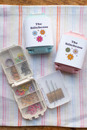 Three small storage cases labeled 'The Stitchcase' arranged on a striped fabric surface. One case is open and shows elastic bands, thread, and pins inside; the other two are closed with pastel lids in mint, pink, and light blue.