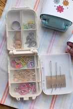 Load image into Gallery viewer, An open white plastic organizer case filled with colorful rubber bands, safety pins, and small sewing or craft accessories, placed on a striped fabric surface next to a floral-patterned container.