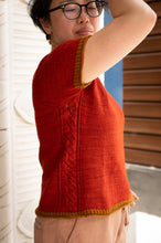 Load image into Gallery viewer, A person wearing a short-sleeve knit top in a burnt orange-red color with mustard yellow ribbed trim at the sleeve and hem, paired with beige pants. The person is shown from a side/back angle against a background of white textured wall and brown wooden shutters.