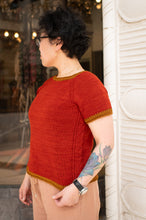 Load image into Gallery viewer, A woman wearing a short-sleeve knit top in rust red with mustard yellow trim, standing outdoors next to a white building.