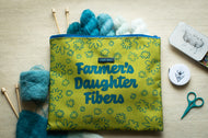 Green pouch with 'Farmer's Daughter Fibers' branding, surrounded by yarn and knitting tools on a wooden surface.