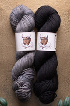 Load image into Gallery viewer, Two skeins of yarn, one gray and one black, with labels on a beige background.