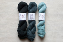 Load image into Gallery viewer, Three skeins of yarn in black, gray, and blue on a beige surface with labels.