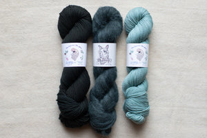 Three skeins of yarn in black, gray, and blue on a beige surface with labels.